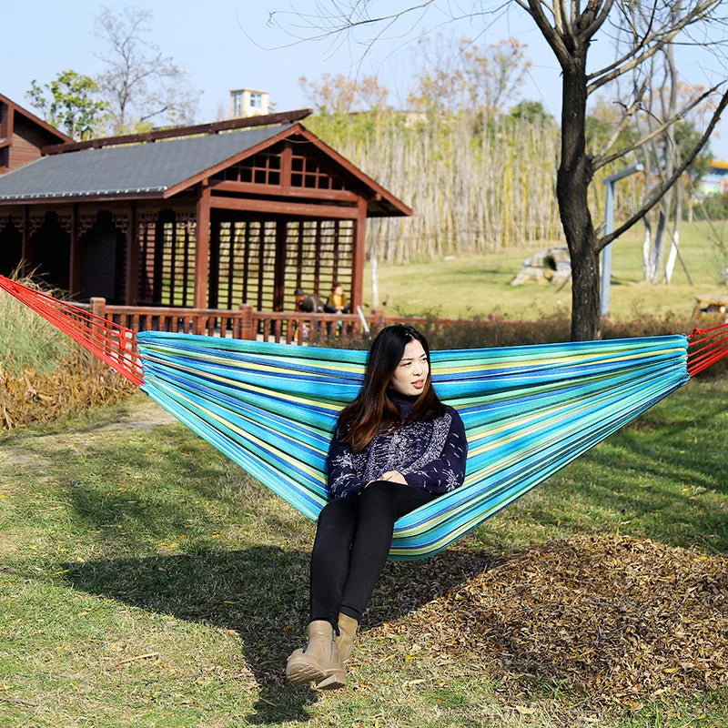 Woman sitting in a colorful striped hammock outdoors with a wooden cabin in the background.