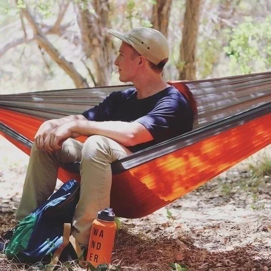 Man sitting in a red hammock in a forest with a water bottle labeled 'Wander' on the ground.