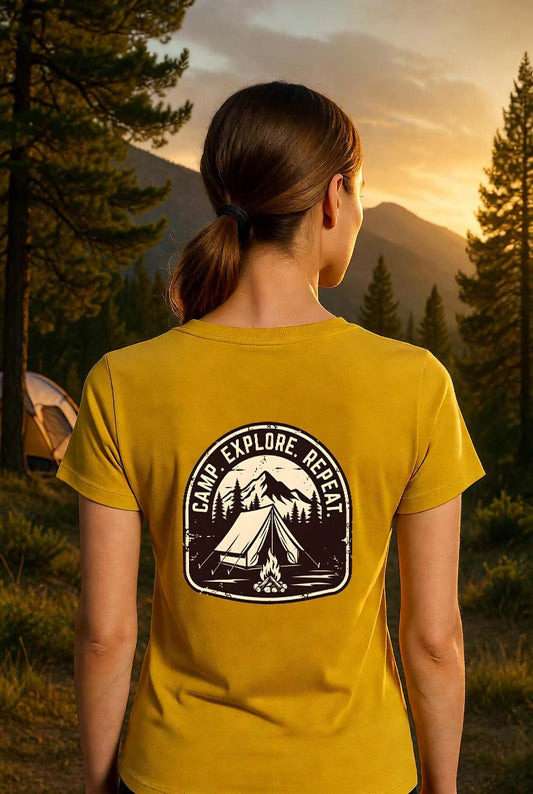 Rear view of a woman wearing a mustard yellow CAMP. EXPLORE. REPEAT. vintage camping t-shirt in a pine forest camping scene at golden hour