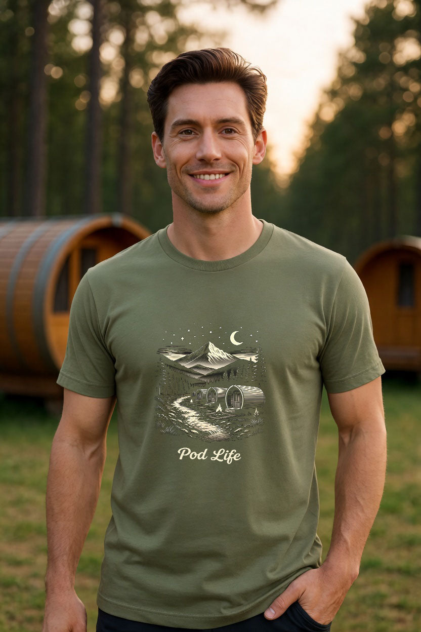 Handsome man wearing a plain sage green organic t-shirt in a beautiful woodland glamping setting with barrel pods in the background at golden hour.