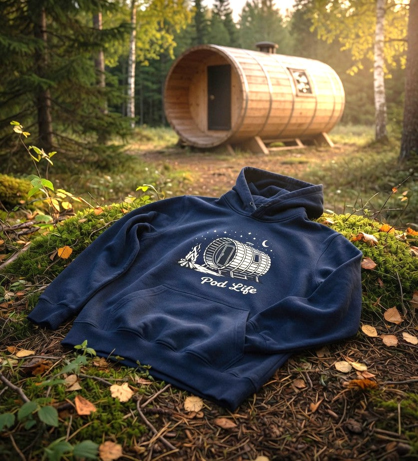 Navy blue Pod Life hoodie with barrel pod, campfire and moon graphic draped naturally on a mossy forest floor with a real wooden glamping pod in the golden hour background.